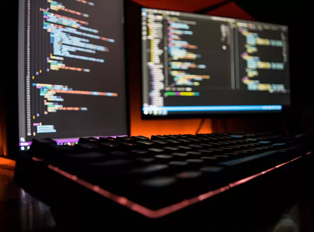 Foto de um teclado e dois monitores exibindo programação avançada em suas telas