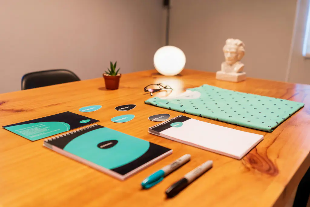 Vista de cima de mesa de designer com prancha de identidade visual, contendo esboços de logotipo, amostras de paleta de cores, guias tipográficos e mockups em papel e tablet, em ambiente minimalista com luz natural suave